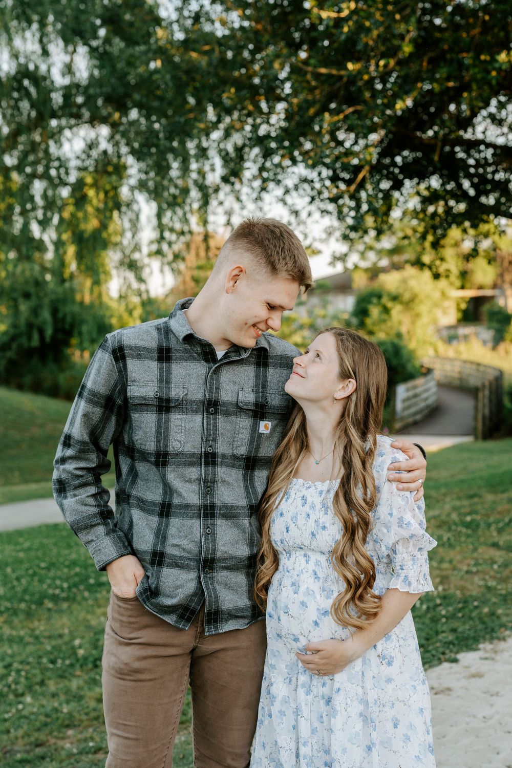 A young couple shares a tender moment outdoors, with the woman in a floral dress cradling her baby bump.