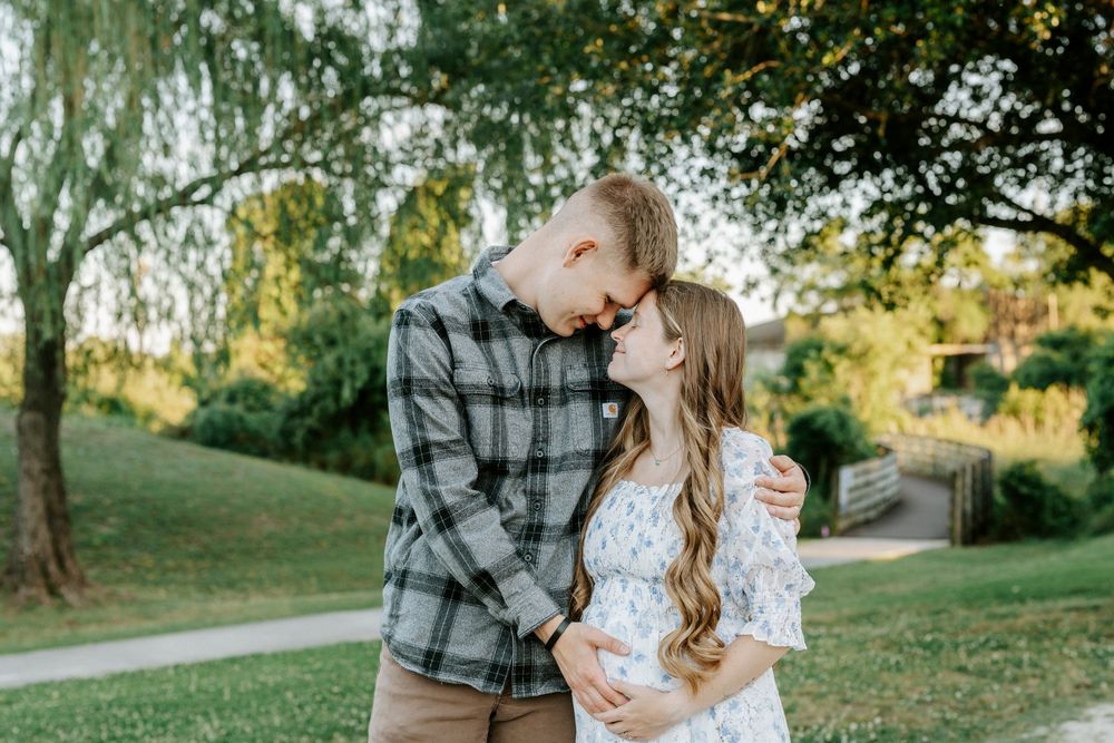 A couple shares a tender moment outdoors, the woman in a white lace dress cradling her baby bump during a maternity photo session.