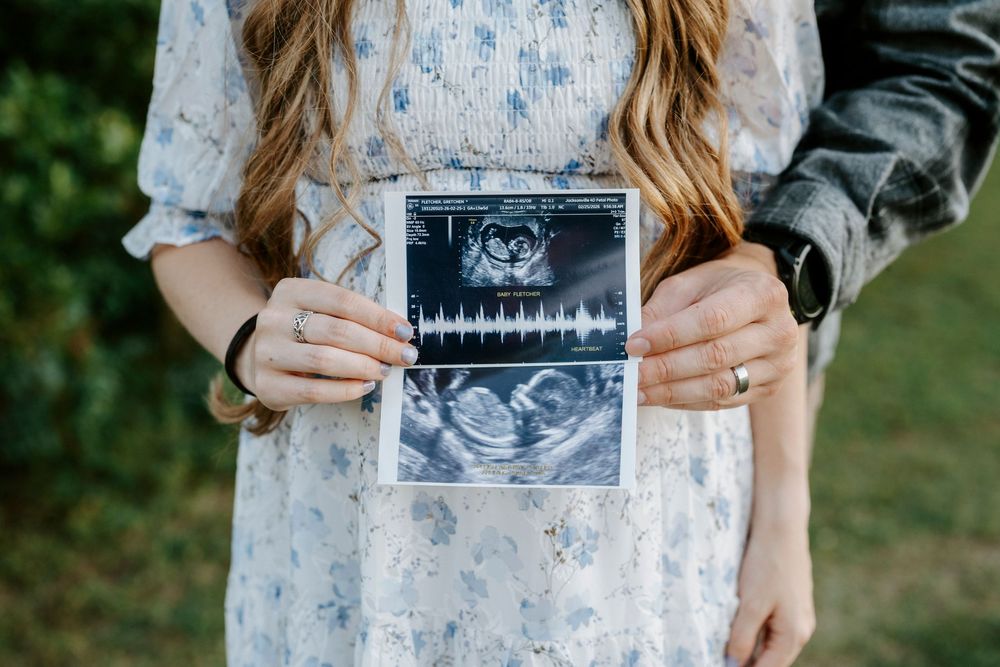 Pregnant couple holding ultrasound sonogram photo outdoors, woman in floral dress showing baby scan image.