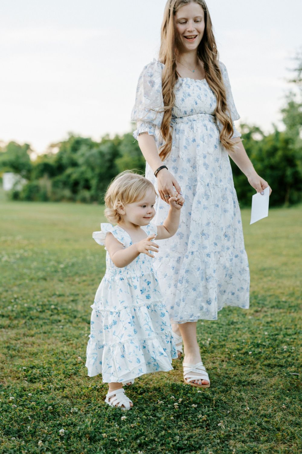 Mother and toddler daughter in matching floral dresses holding hands while walking on green grass outdoors.
