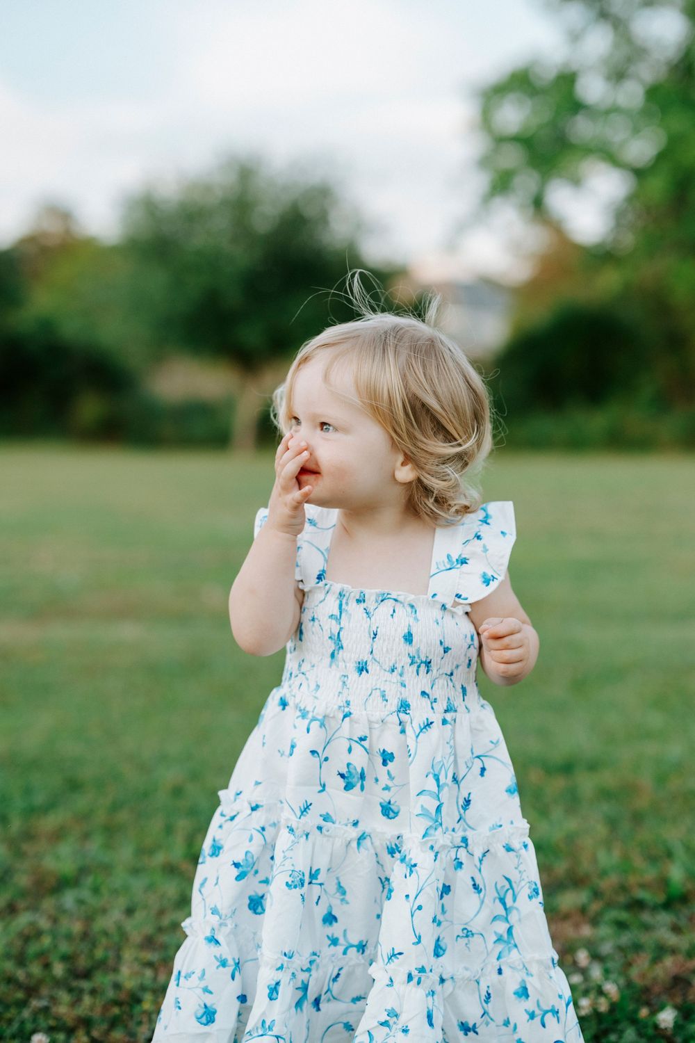Toddler girl in white floral dress standing on green grass, touching her nose, with trees in background.