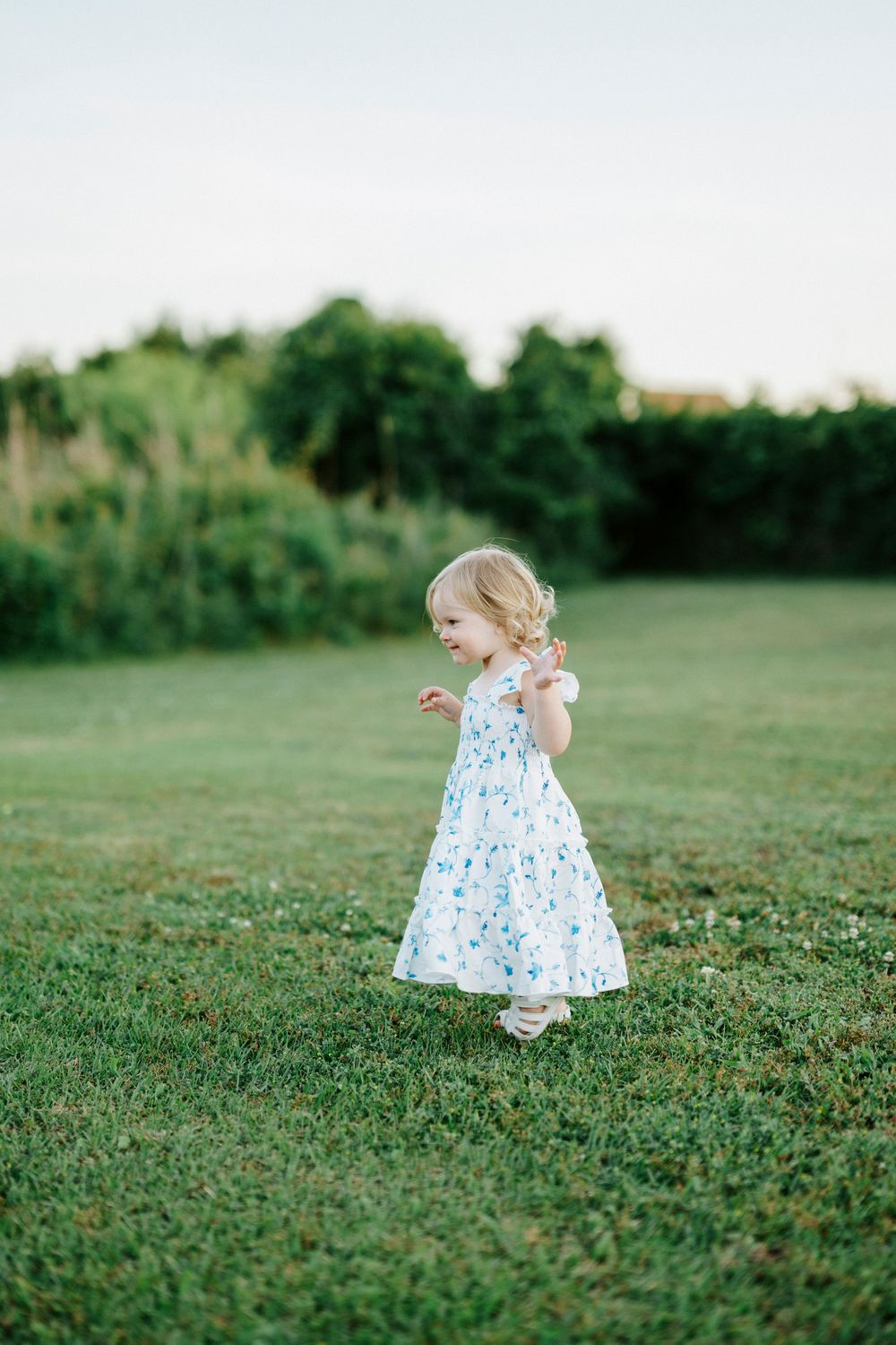 Toddler girl in white floral dress playing on green grass lawn outdoors during golden hour.