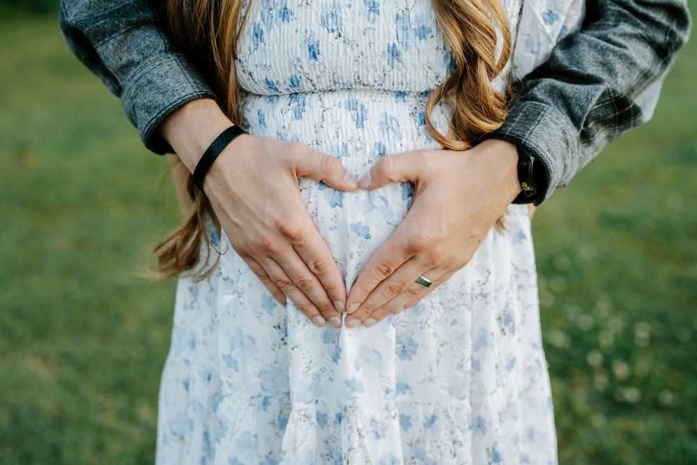 Couple forming heart shape with hands on pregnant belly, woman in floral dress outdoors.