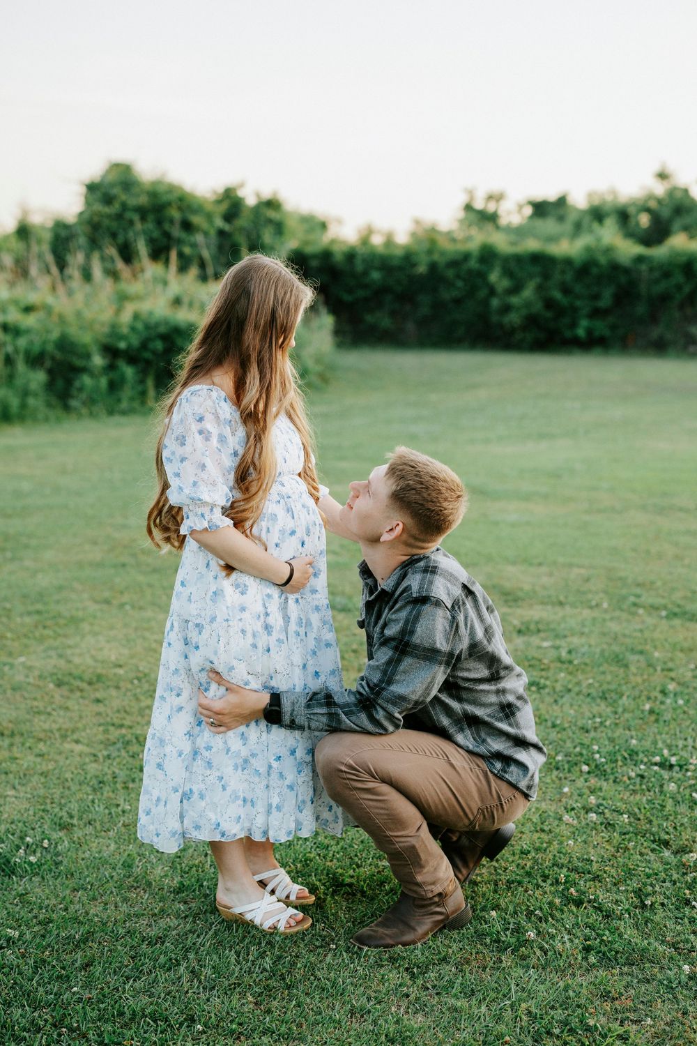 A man kneels on green grass kissing a pregnant woman's belly during an outdoor maternity photo session at sunset.