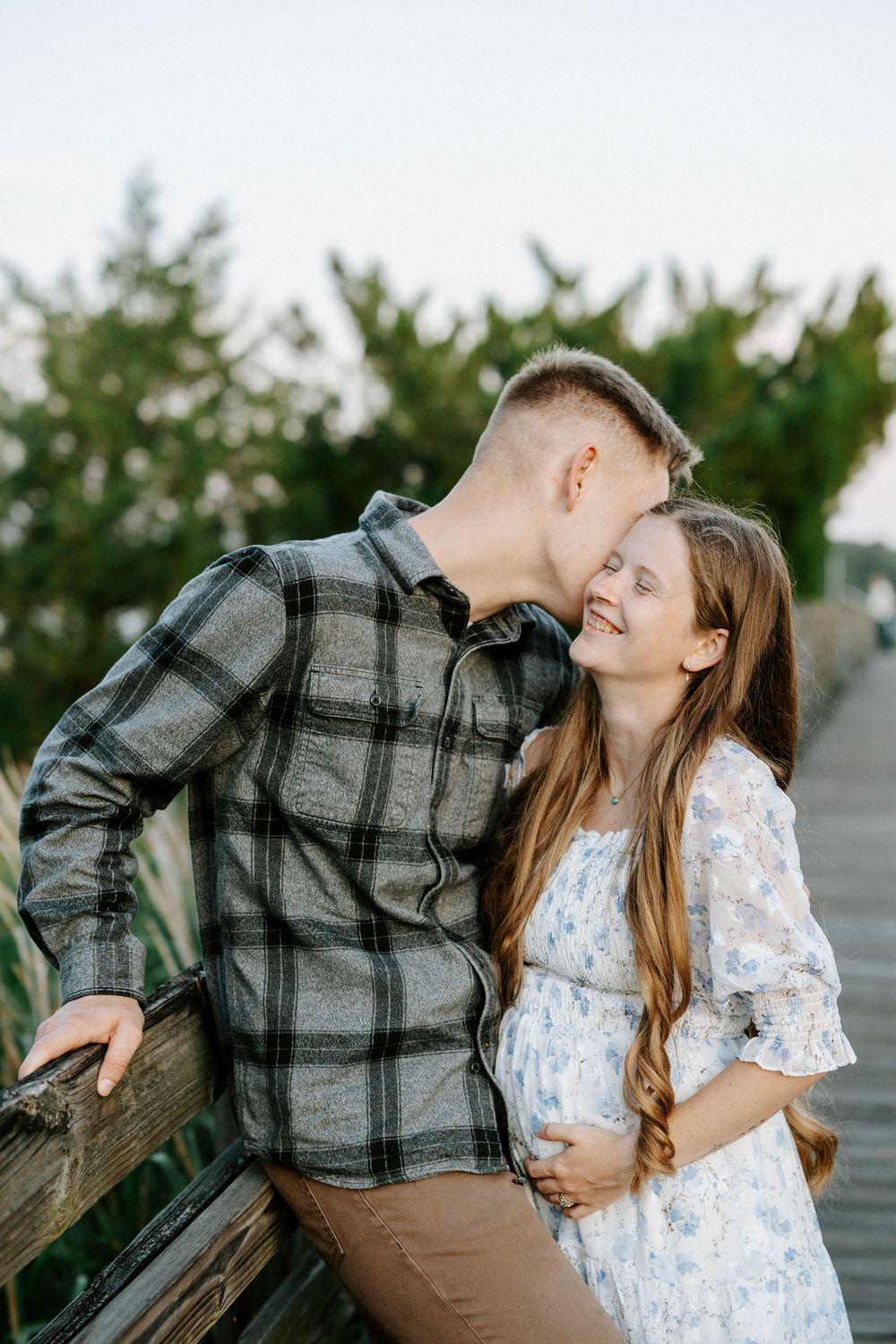 A man kisses a smiling pregnant woman on a wooden boardwalk surrounded by greenery.