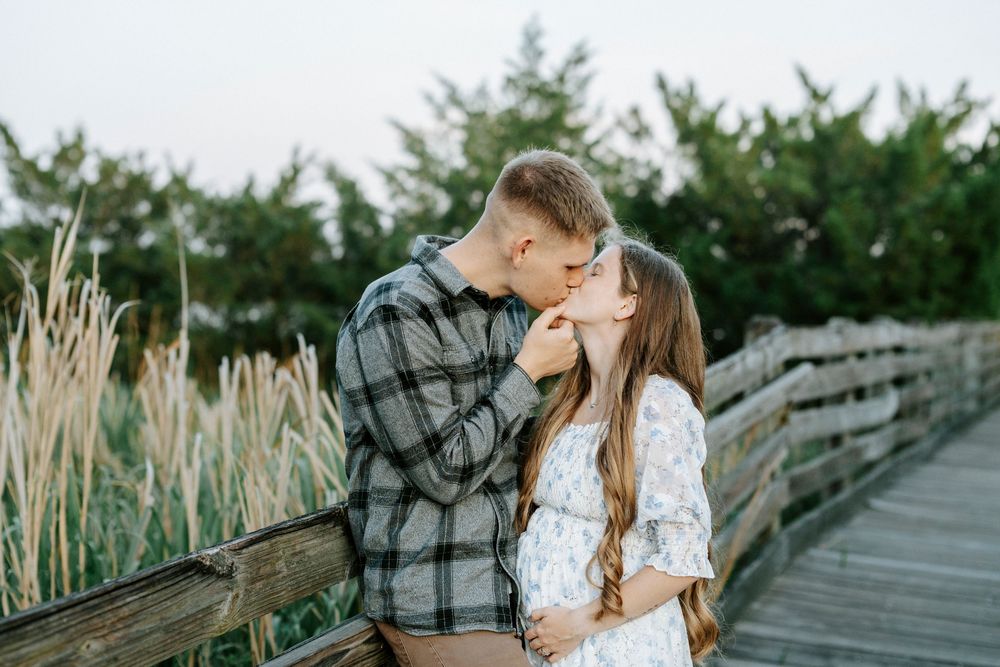 Couple sharing a romantic kiss on a wooden boardwalk surrounded by tall grass and green trees at sunset.