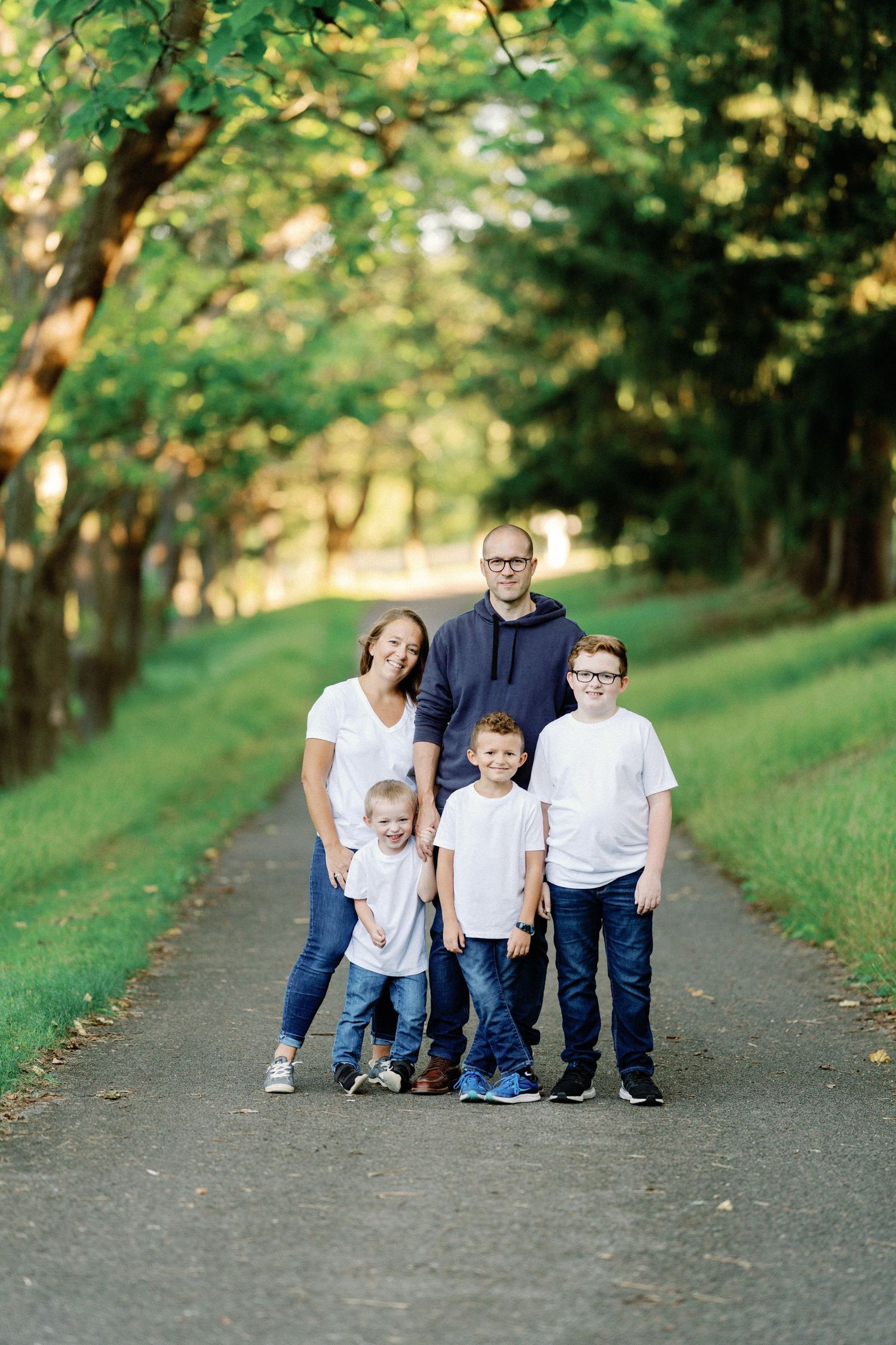 Family of five posing on a tree-lined path outdoors, parents with three young boys in casual white and blue outfits.