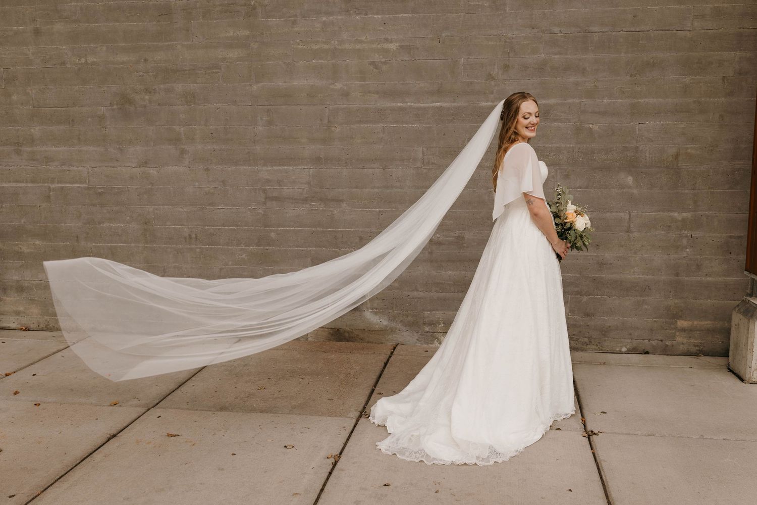 Bride in a flowing white gown with a long veil blowing in the wind outdoors.