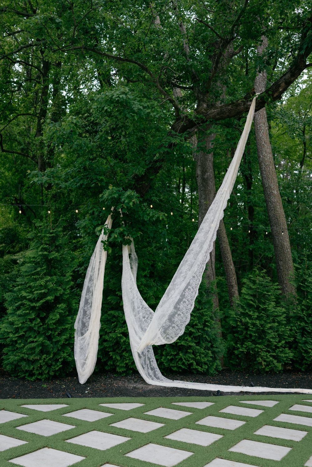 White lace fabric draped between trees in an outdoor wedding ceremony space with string lights and lush green trees.