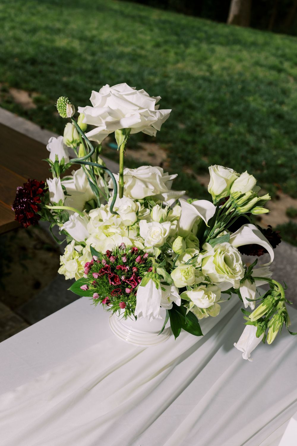 Elegant white floral arrangement with roses, calla lilies, and red accents in a white vase on a draped table outdoors.