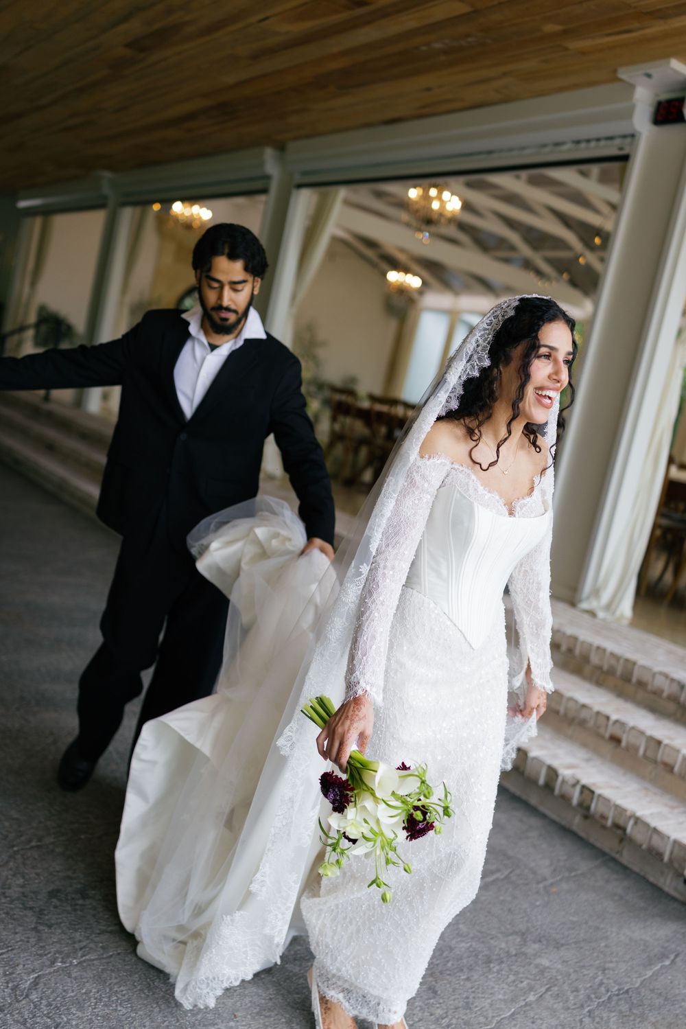 Bride in lace wedding gown holding bouquet walks with groom in black suit through elegant venue corridor.