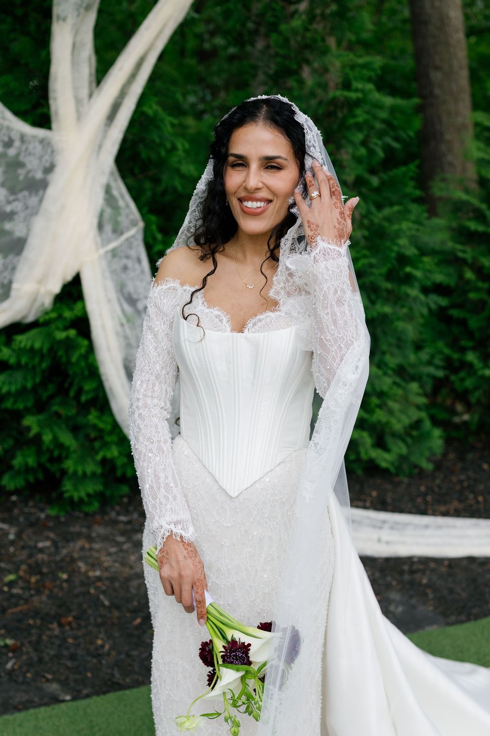Smiling bride in white lace wedding gown with veil, holding dark floral bouquet outdoors among green trees.