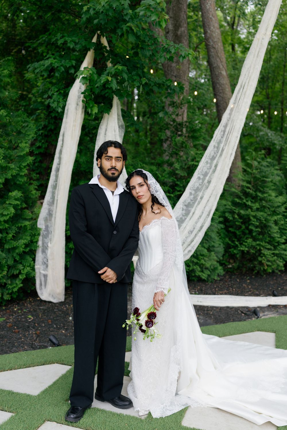 Bride and groom posing outdoors at elegant garden wedding ceremony with white draping and lush green trees.