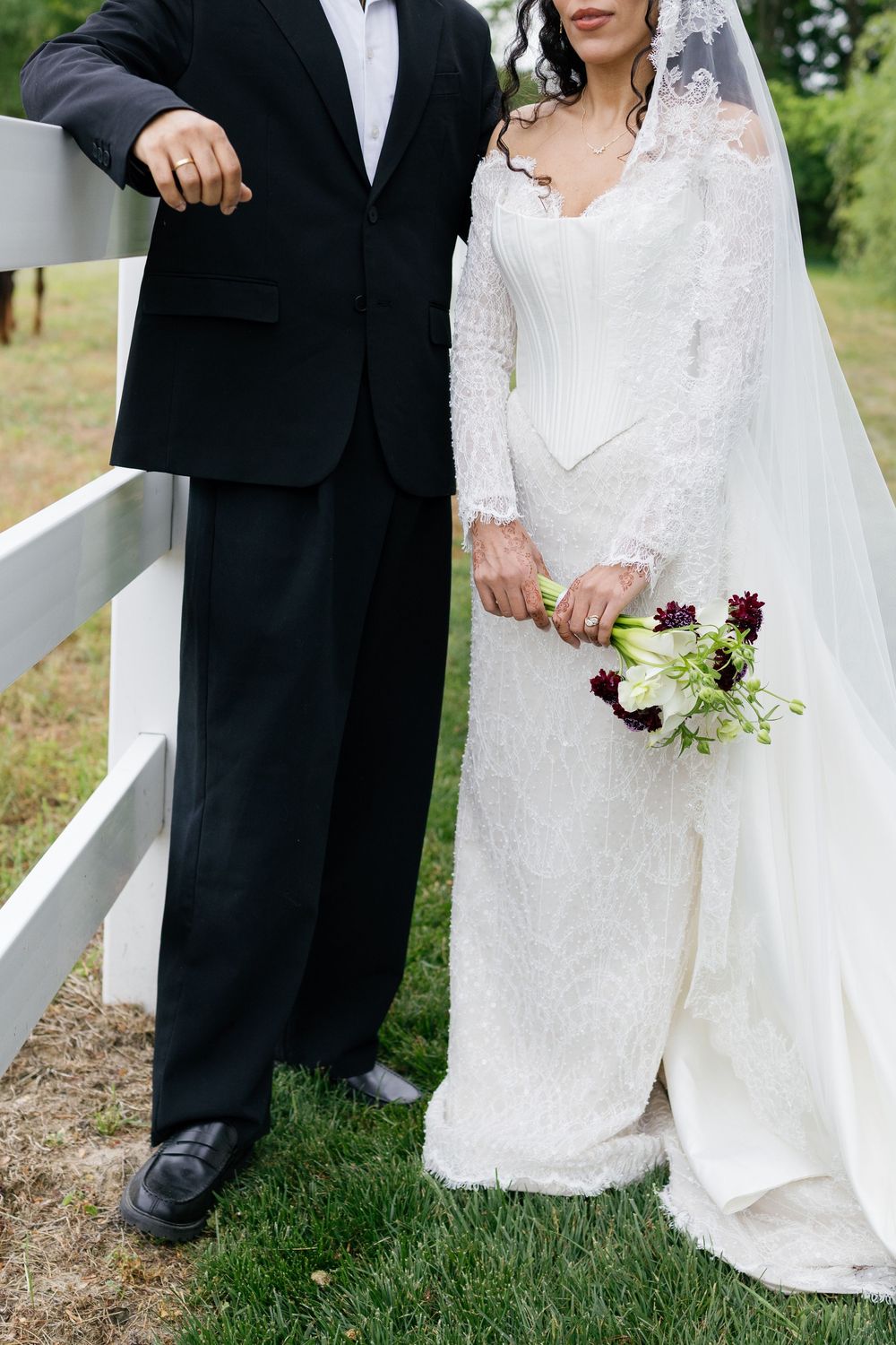 Bride in lace gown holding dark floral bouquet stands with groom in black suit near white fence outdoors.