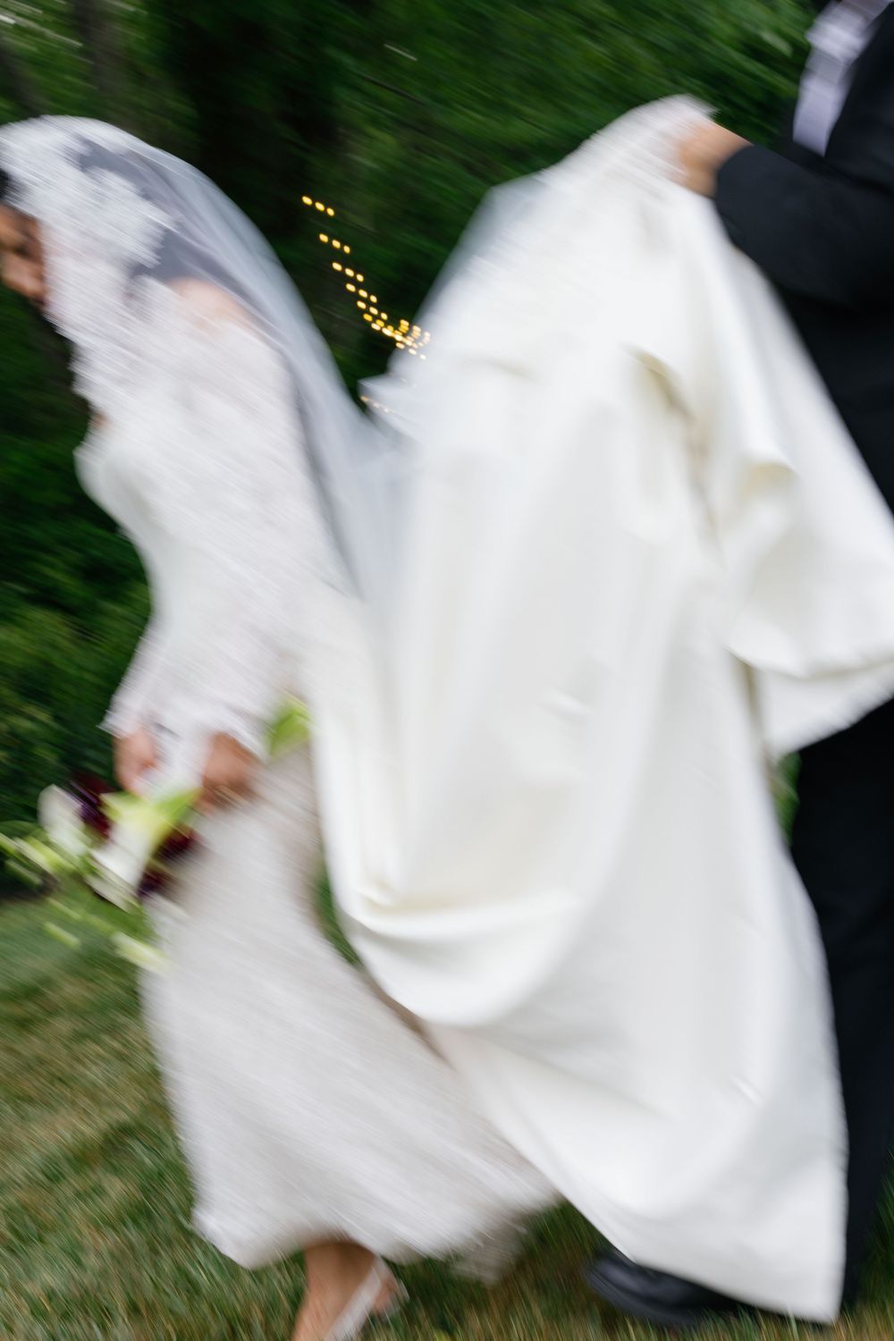 Blurred motion shot of a bride in white gown and veil holding bouquet, walking with groom outdoors.
