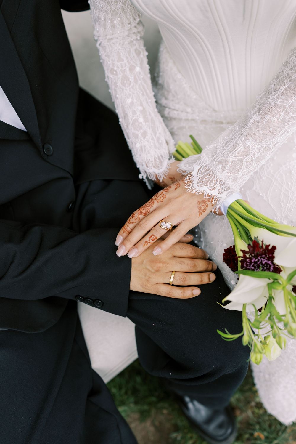 Bride and groom hands with wedding rings and henna, holding a bouquet of white and burgundy flowers.