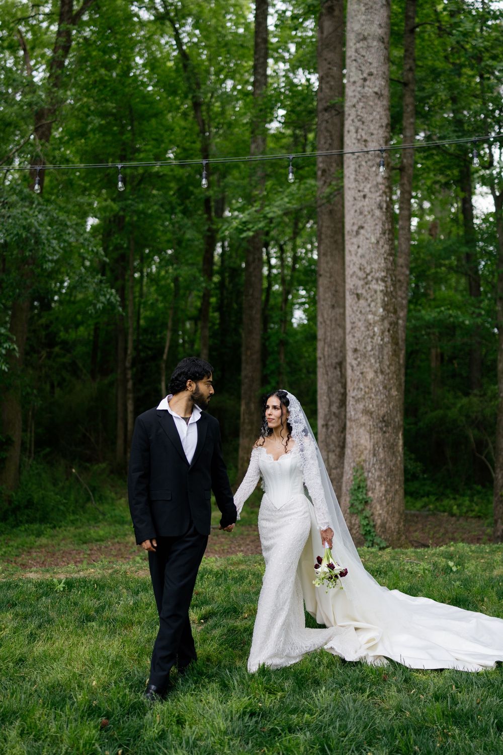 Bride and groom hold hands walking in a lush green forest, she in a white gown, he in a black suit.