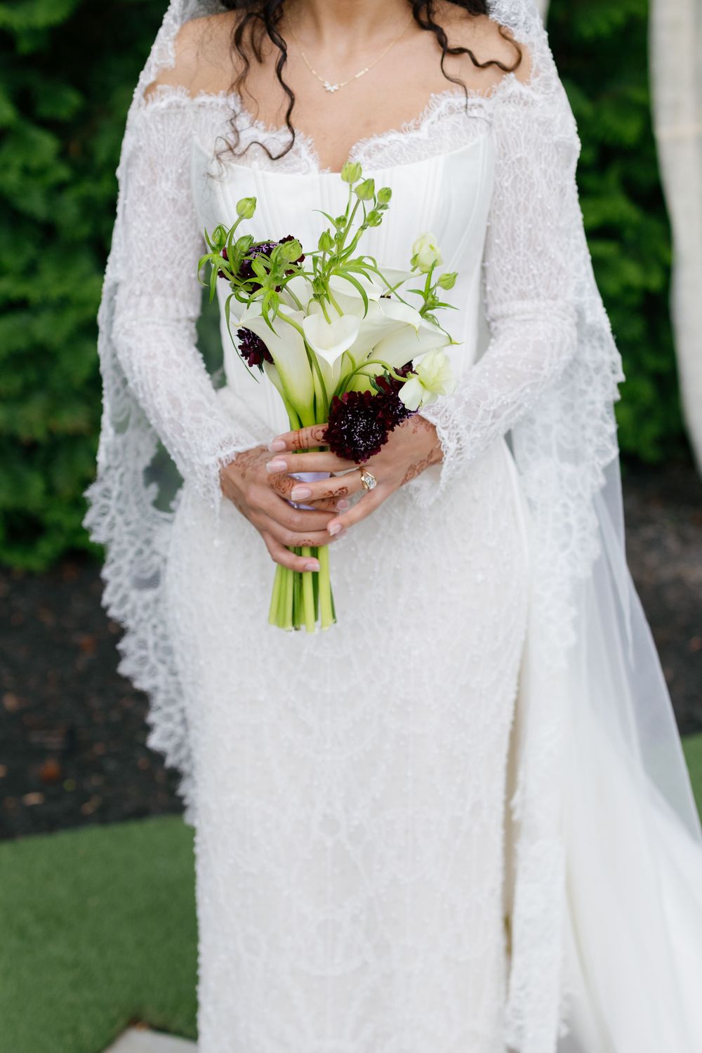 Bride in lace wedding gown and veil holding elegant bouquet of white calla lilies and dark burgundy flowers.
