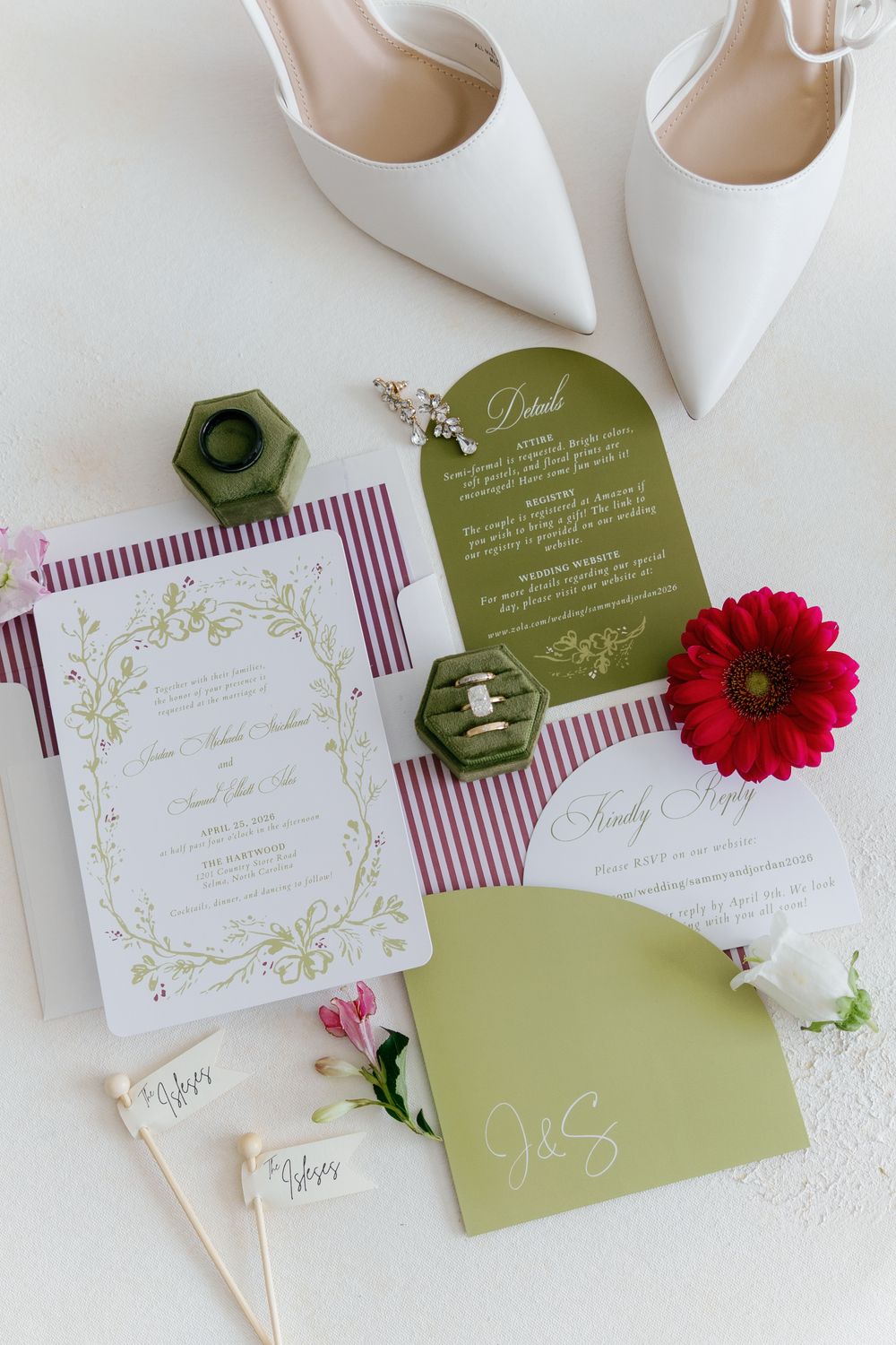 Elegant wedding invitation suite with green and burgundy accents, ring box, red flower, and white heels on white background.
