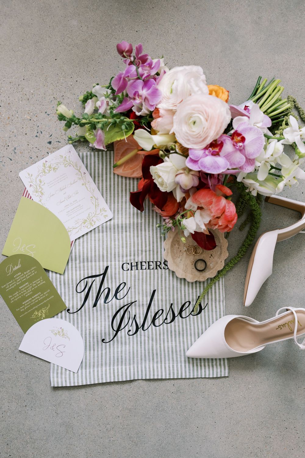 Elegant wedding flat lay featuring a colorful bridal bouquet, white heels, invitations, and a striped cloth reading Cheers The Isleses.