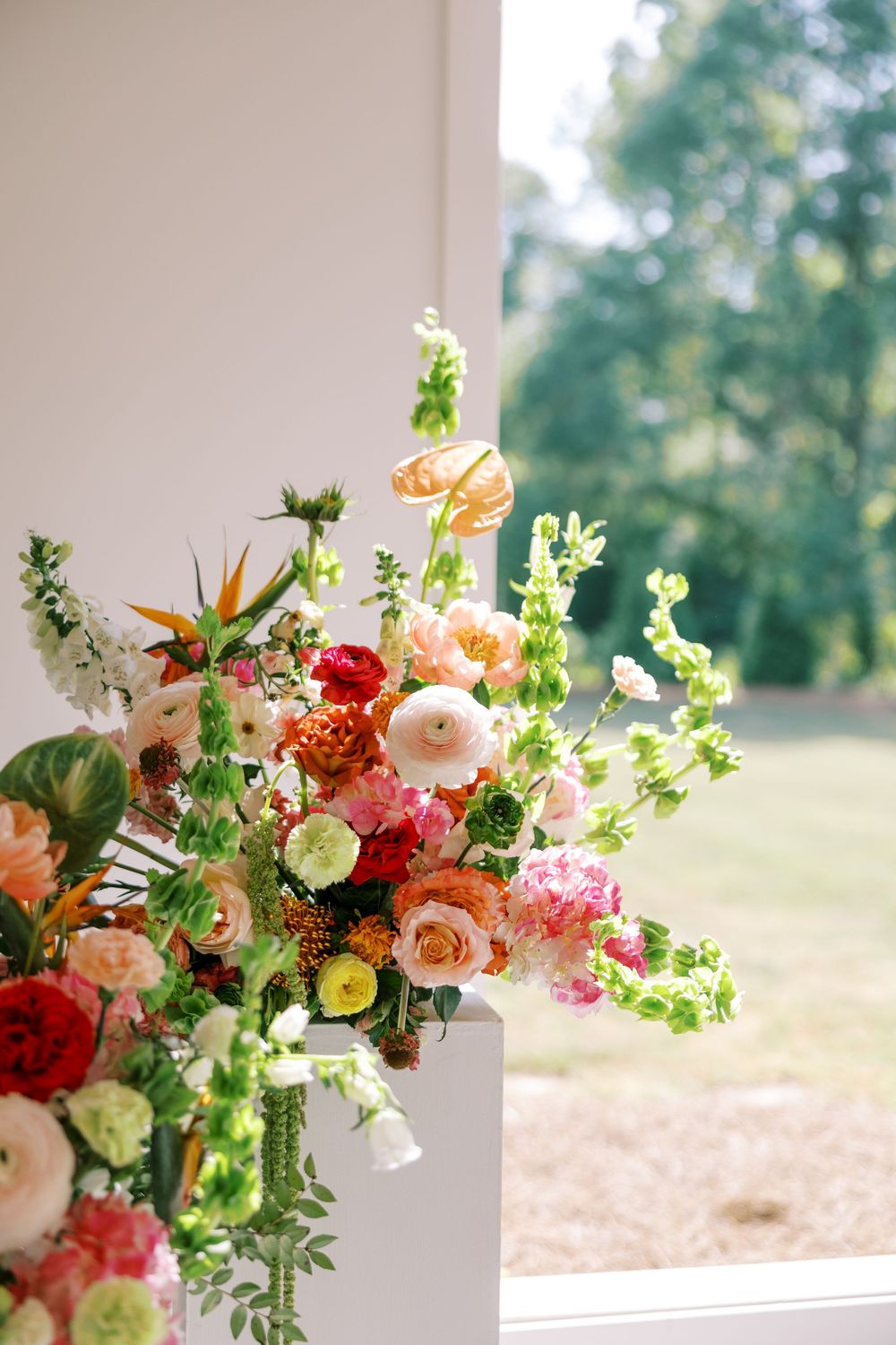 Lush wedding floral arrangement with ranunculus, roses, and bird of paradise blooms displayed on a white pedestal near a sunlit window.