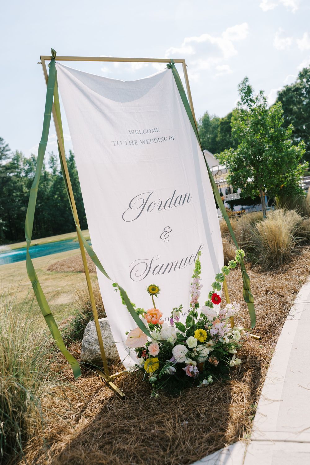 Outdoor wedding welcome sign for Jordan and Samuel on gold stand with colorful floral arrangement and green ribbons.