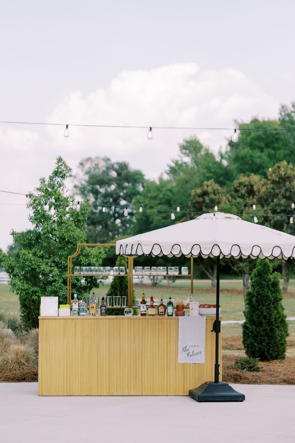 Outdoor wedding bar setup with a yellow bamboo counter, striped umbrella, string lights, and bottles of spirits surrounded by greenery.