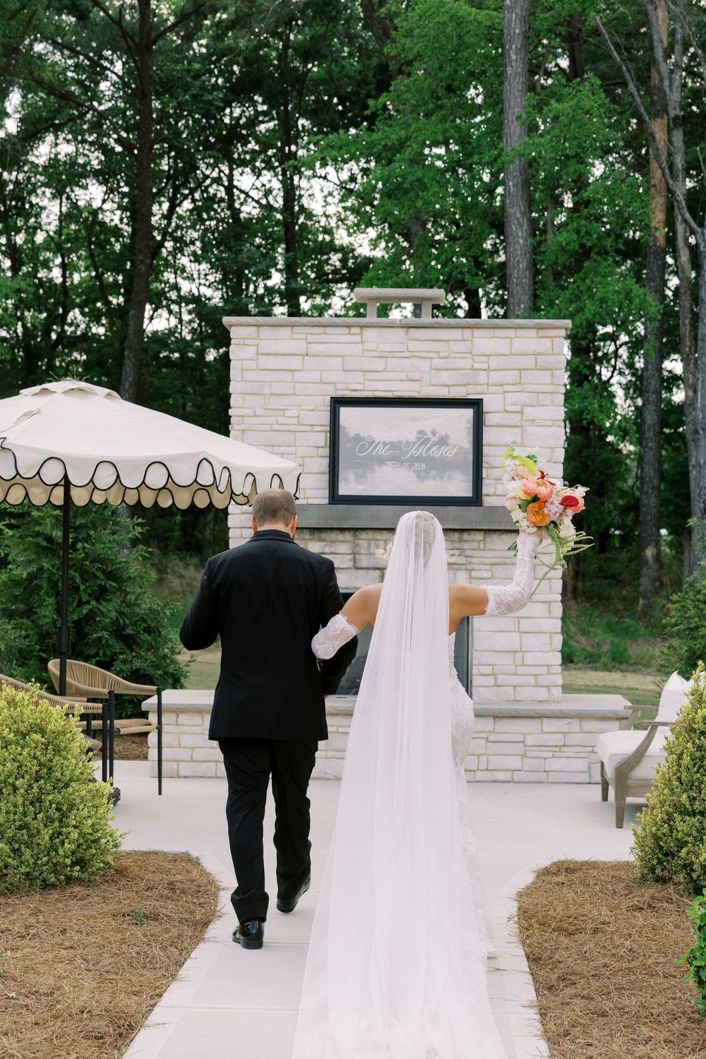 Bride in white veil and groom in black suit walk toward stone fireplace altar at outdoor wedding ceremony.