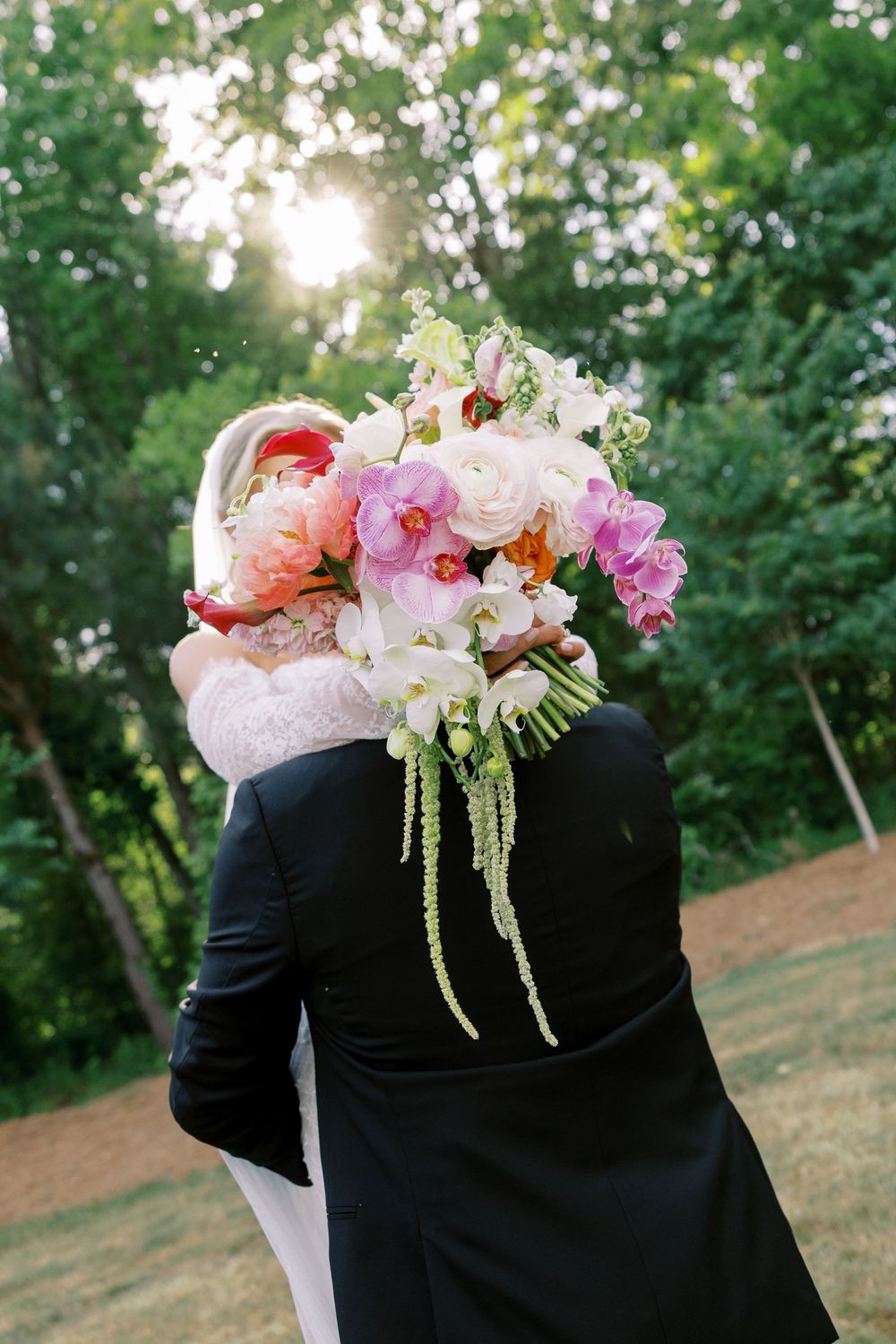 Bride and groom embracing outdoors, faces hidden behind a colorful bouquet of orchids, peonies, and ranunculus in sunlit garden.