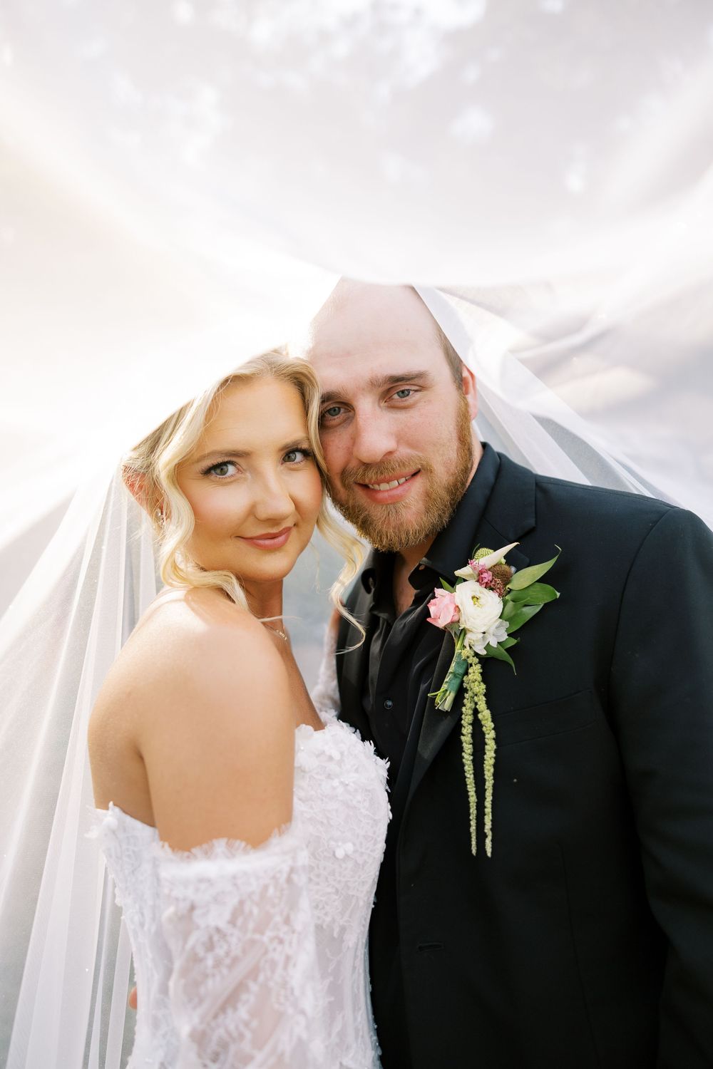 Bride and groom posing under a flowing white veil, smiling at the camera on their wedding day.