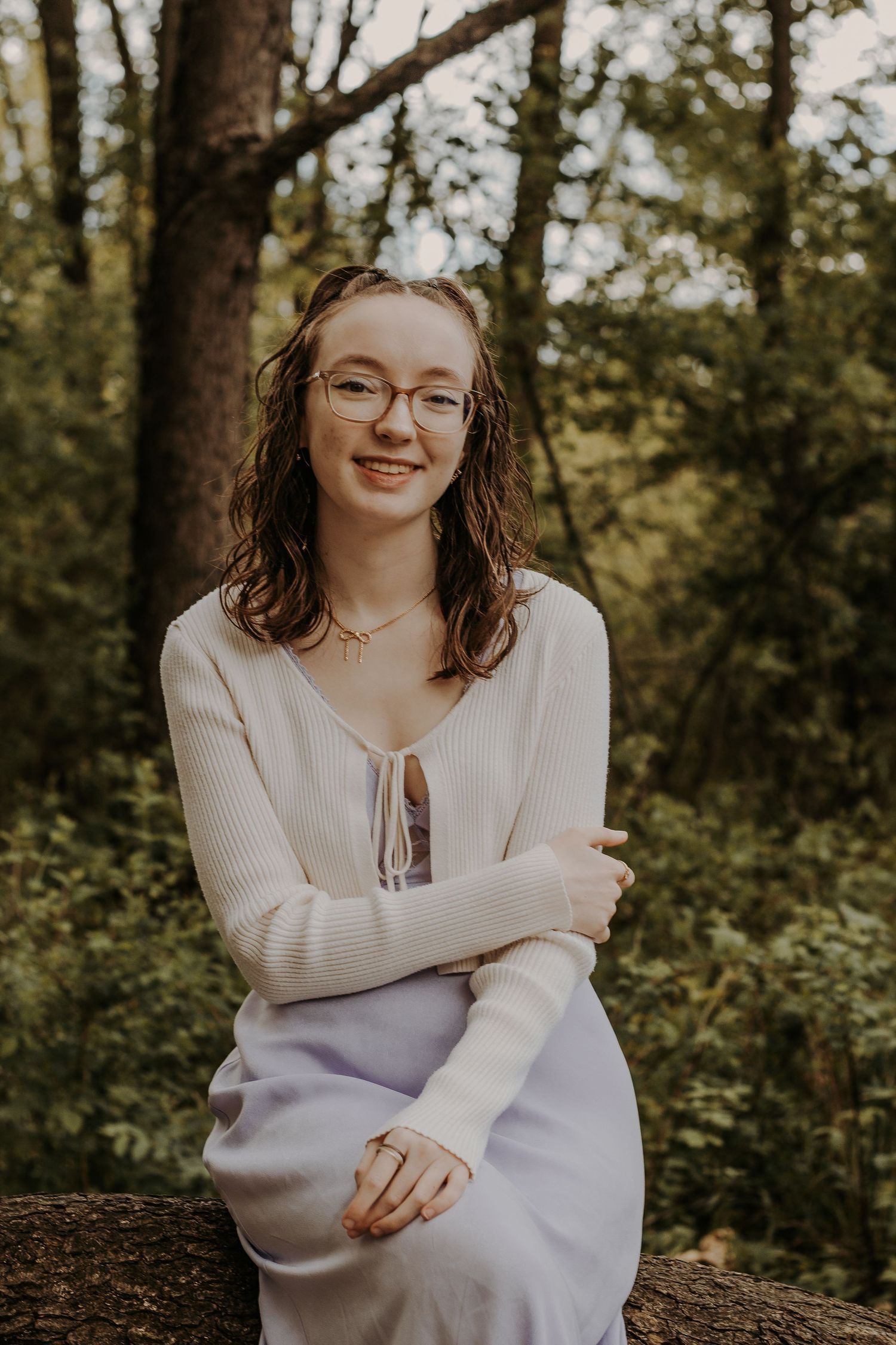 Young woman with glasses smiling in a white sweater and lavender skirt, seated outdoors among lush green trees.