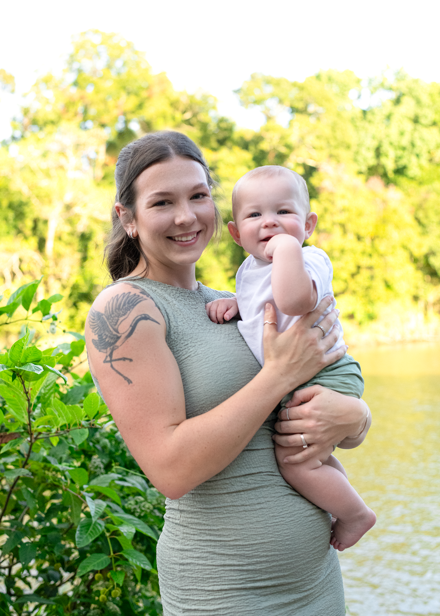 Smiling young mother with tattoo holds happy baby boy near sunlit pond outdoors.
