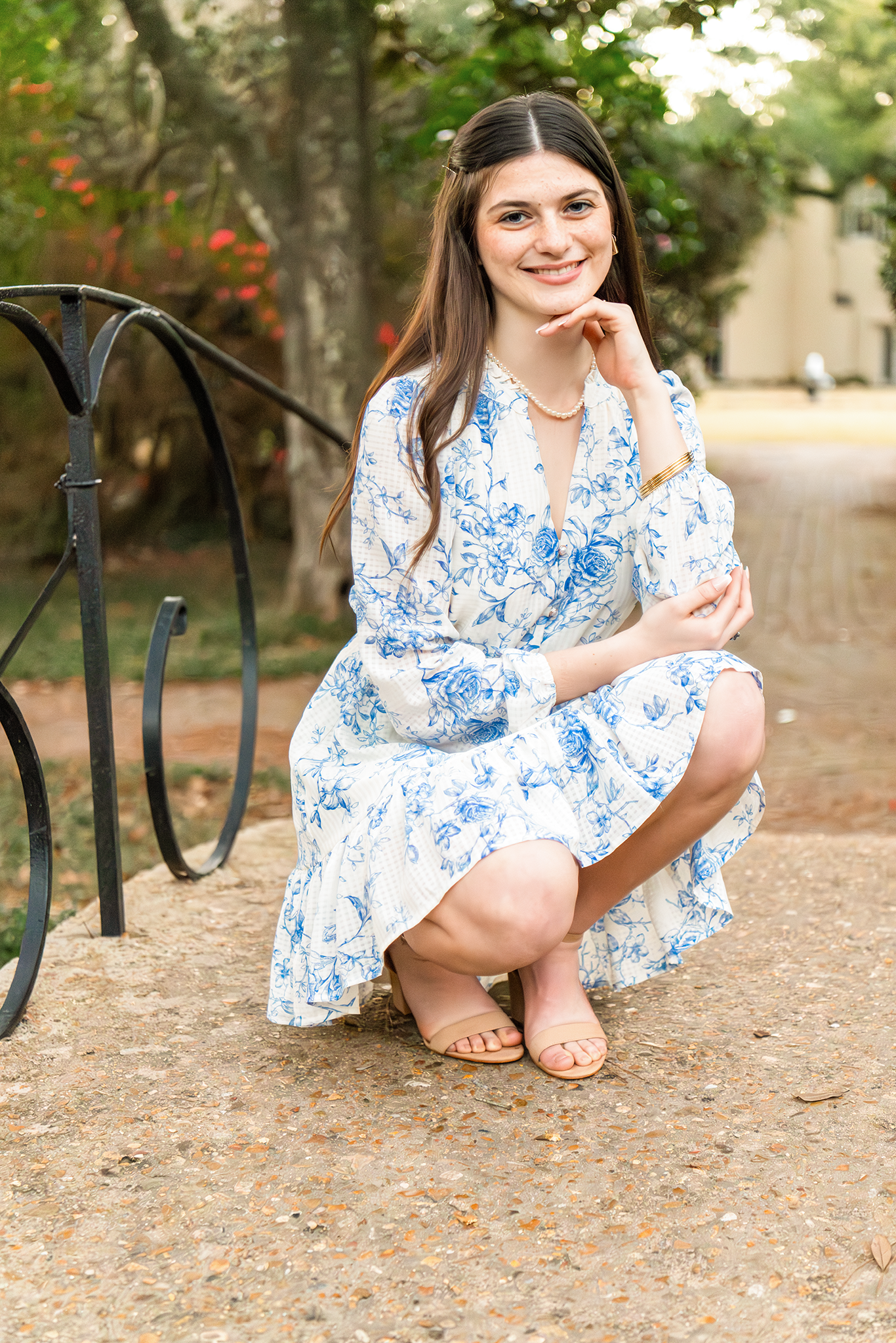 Young woman in a blue floral dress crouches gracefully near a black iron railing outdoors.