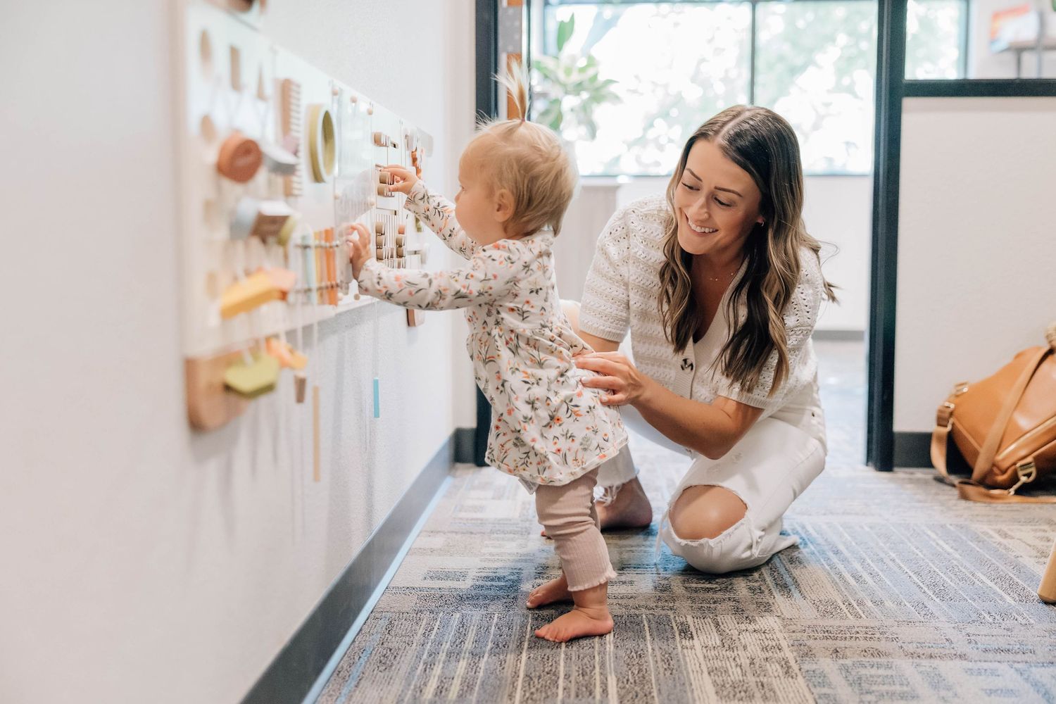 Mother crouching beside toddler exploring colorful sensory wall board in bright modern room.
