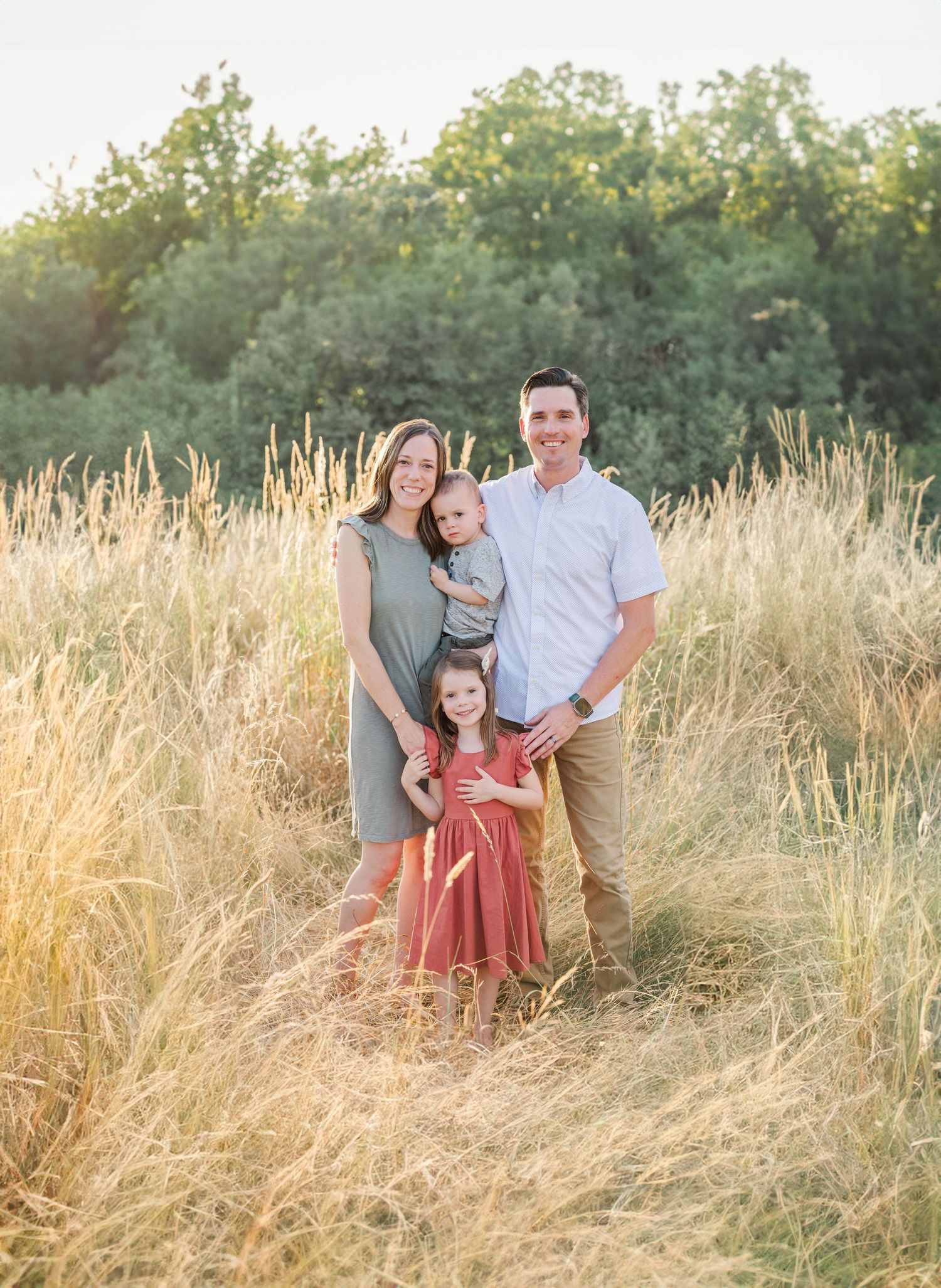 Happy family of four standing in tall golden grass field with lush green trees behind.