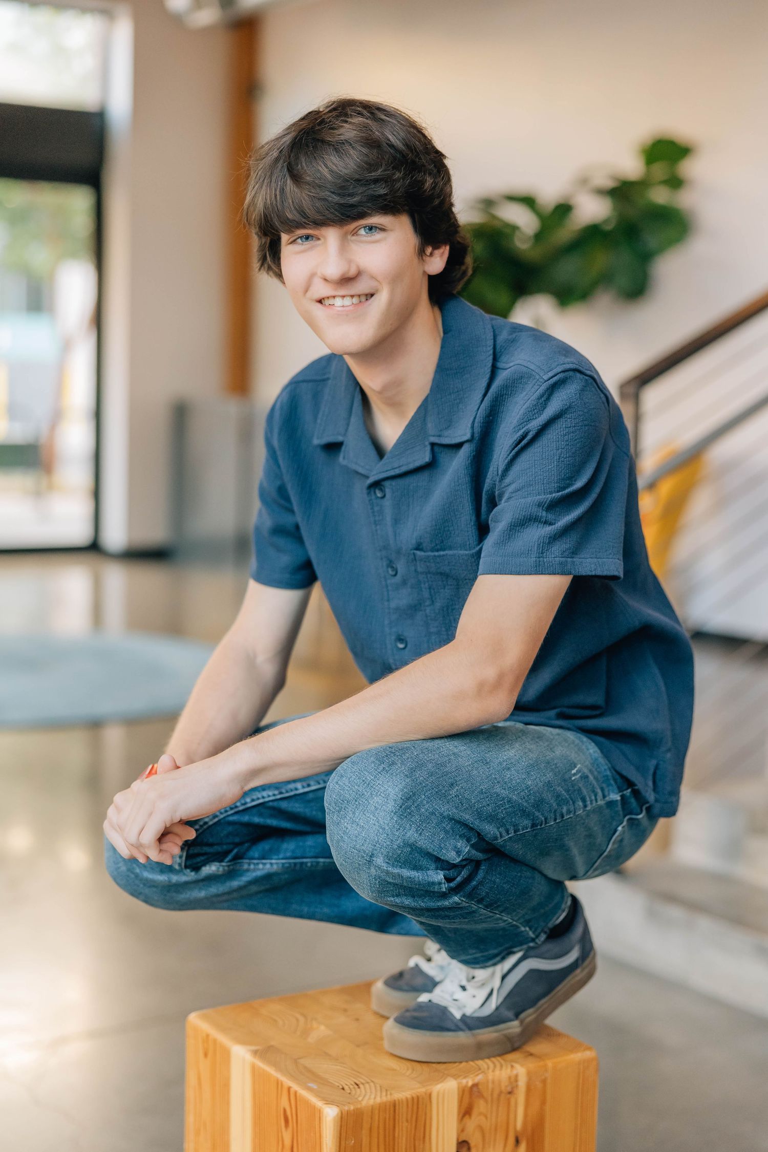 Smiling teenage boy in blue shirt and jeans crouching on a wooden block in a modern indoor setting.