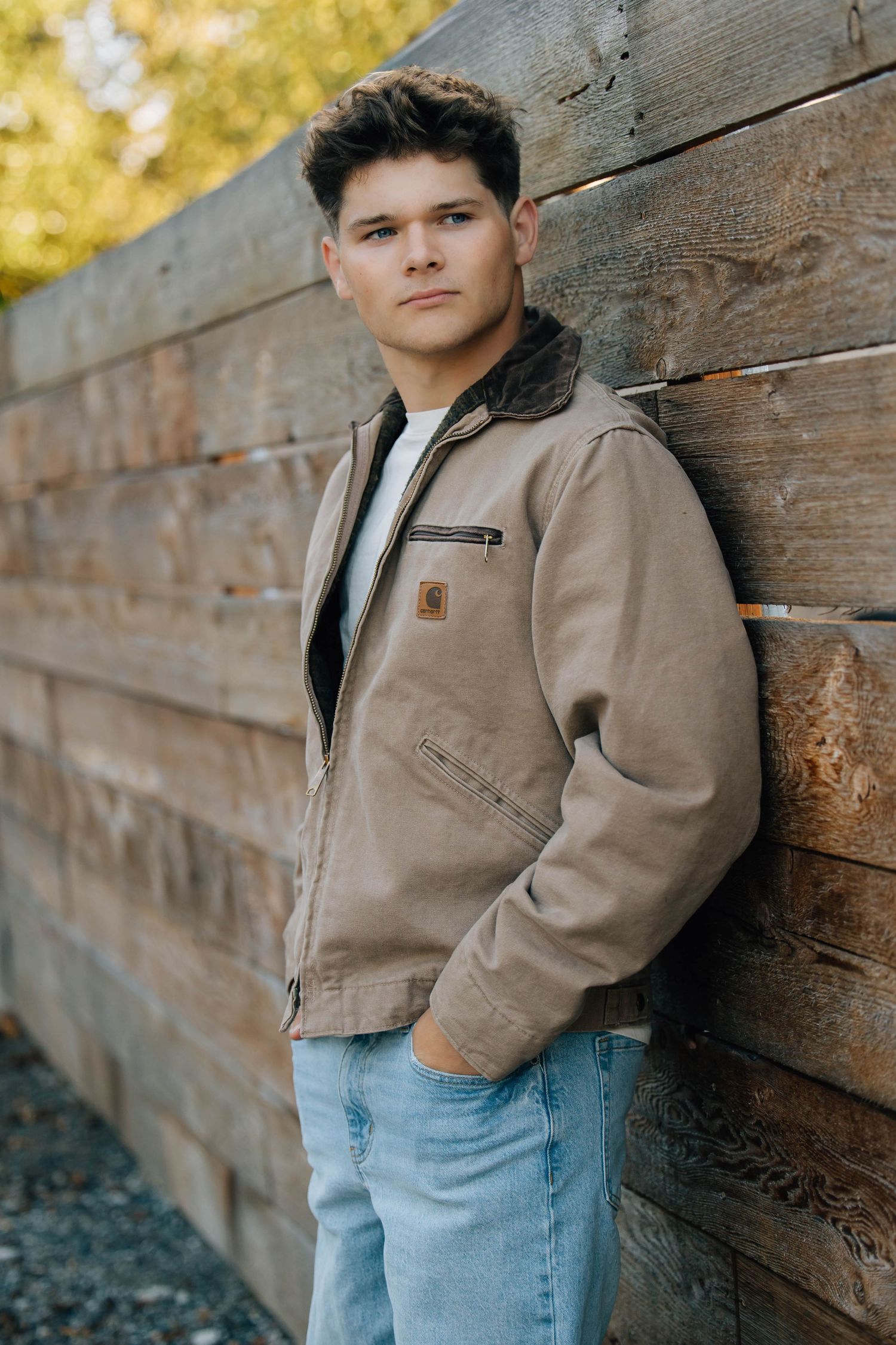 Young man in Carhartt jacket and jeans leaning against rustic wooden wall outdoors.