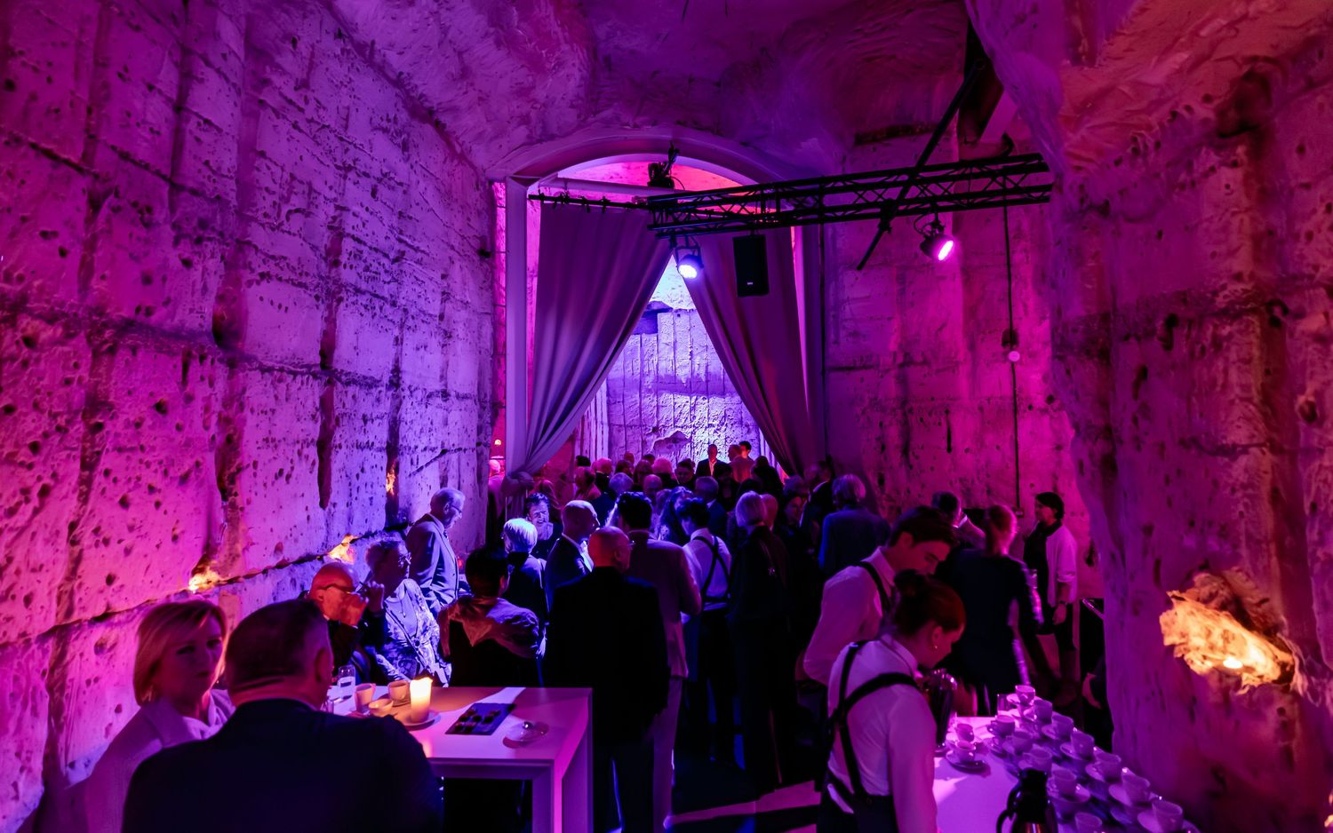 Crowded indoor event venue with purple lighting, stone walls, and elegant draping curtains.