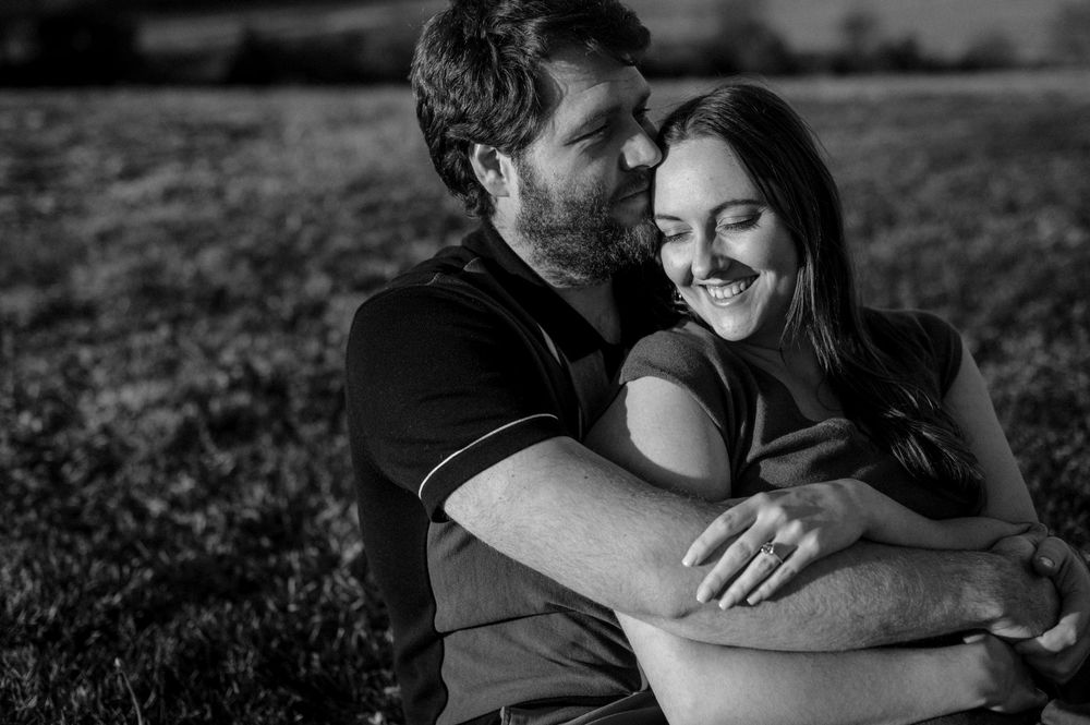 Black and white photo of a couple embracing in a field, woman smiling as man holds her close.