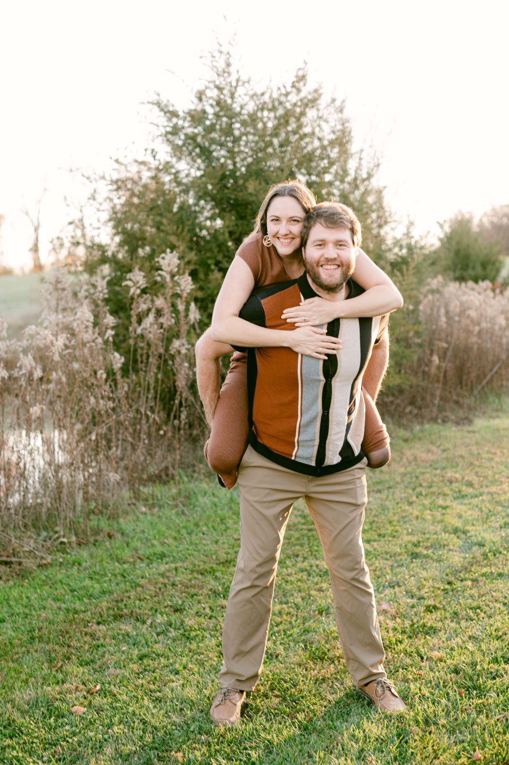 A smiling couple poses outdoors, man giving woman a piggyback ride in a green field with autumn foliage.