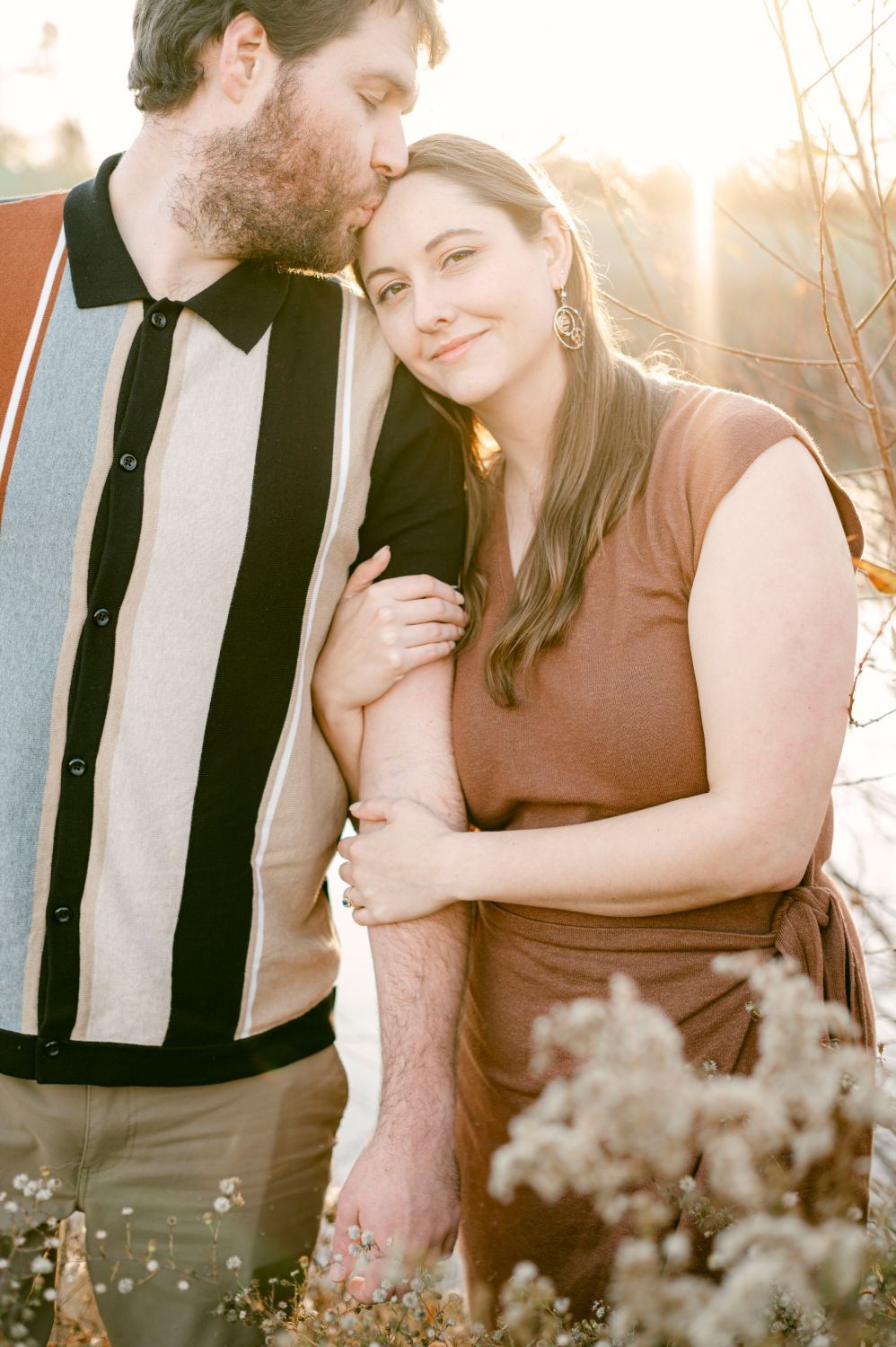 Couple embracing at golden hour, woman leaning into man as he kisses her head near wildflowers.