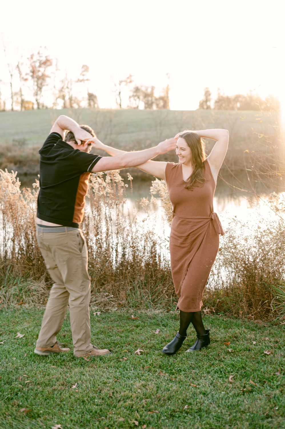 A joyful couple dancing outdoors at golden hour near a serene pond, woman in a brown wrap dress.