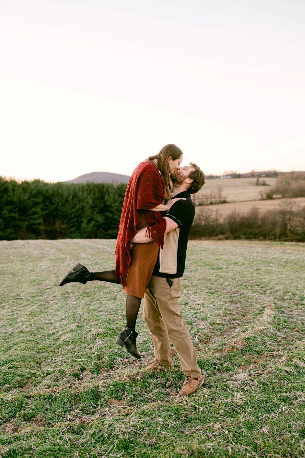 Couple sharing a romantic kiss in an open field, man lifting woman in autumn outfits with rolling hills backdrop.
