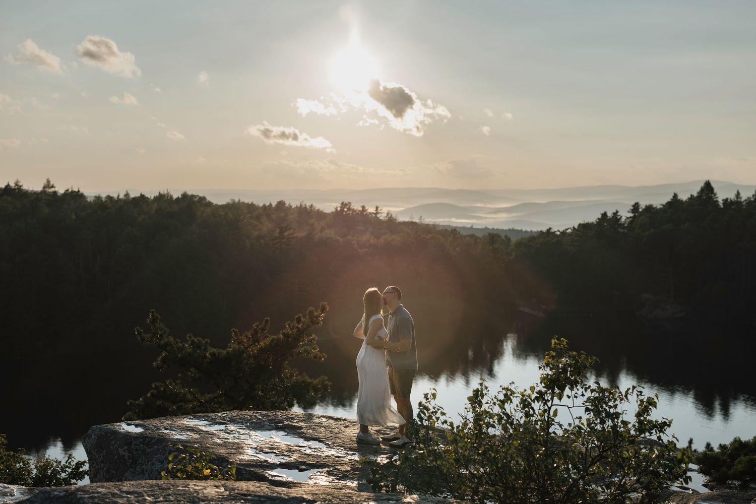 Couple embracing on rocky cliff at sunset with forest and lake backdrop in a stunning outdoor wedding photo.