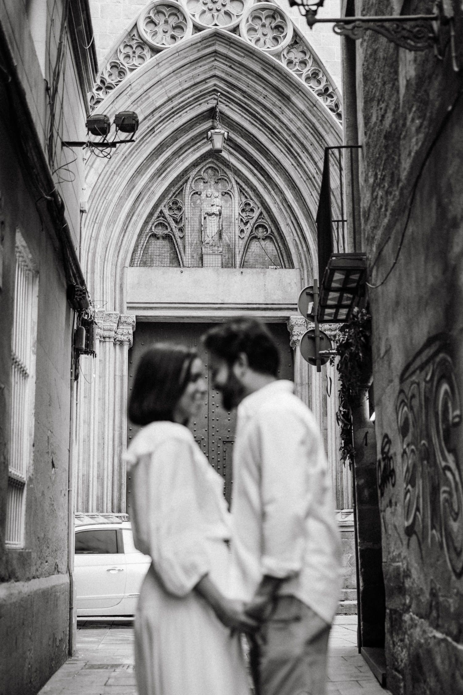 A couple shares a romantic moment under an ornate Gothic archway in a historic European setting.