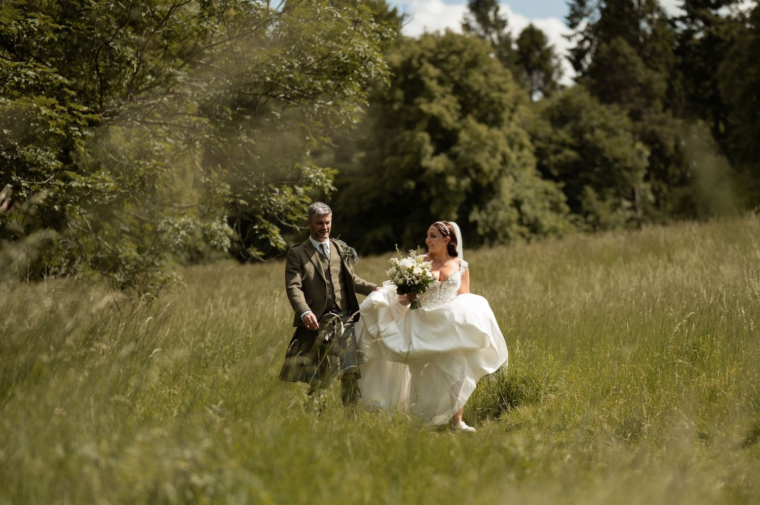 Wedding couple poses for romantic photos in a sunlit meadow with tall grass and trees in the background.