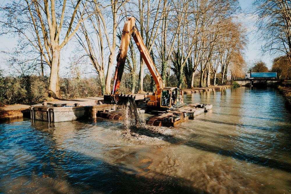 Photographe professionnel Toulouse: Un bateau de dragage en pleine action, bras mécanique plongeant dans l’eau, ciel dégagé au-dessus.