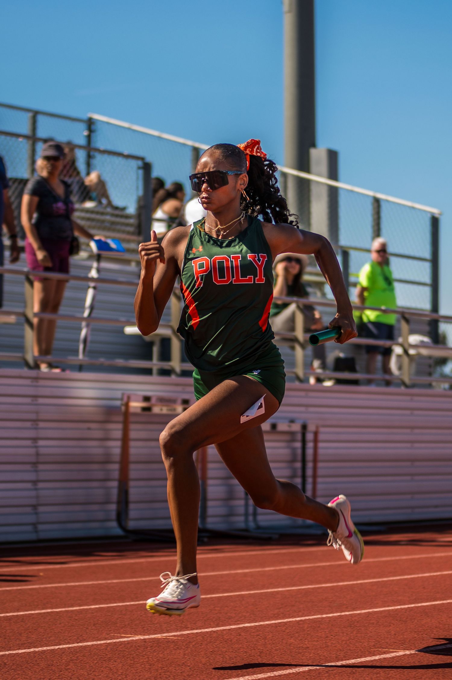 A track athlete wearing a Poly uniform runs on an outdoor track during a sunny day with bleachers visible in background.