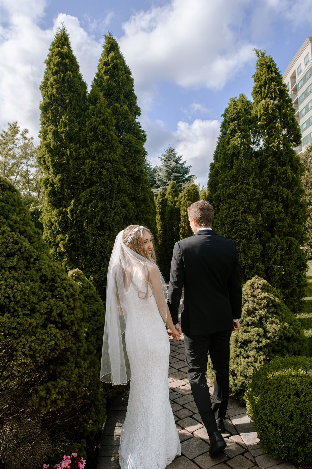 A bride and groom walk along a garden path lined with tall evergreen trees on their wedding day.