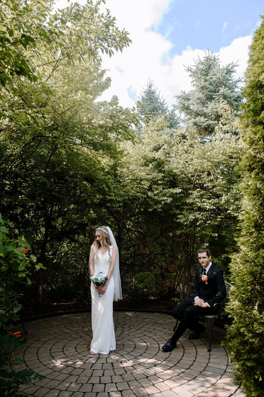 A couple poses in a circular garden setting surrounded by lush green trees and stone pathways.